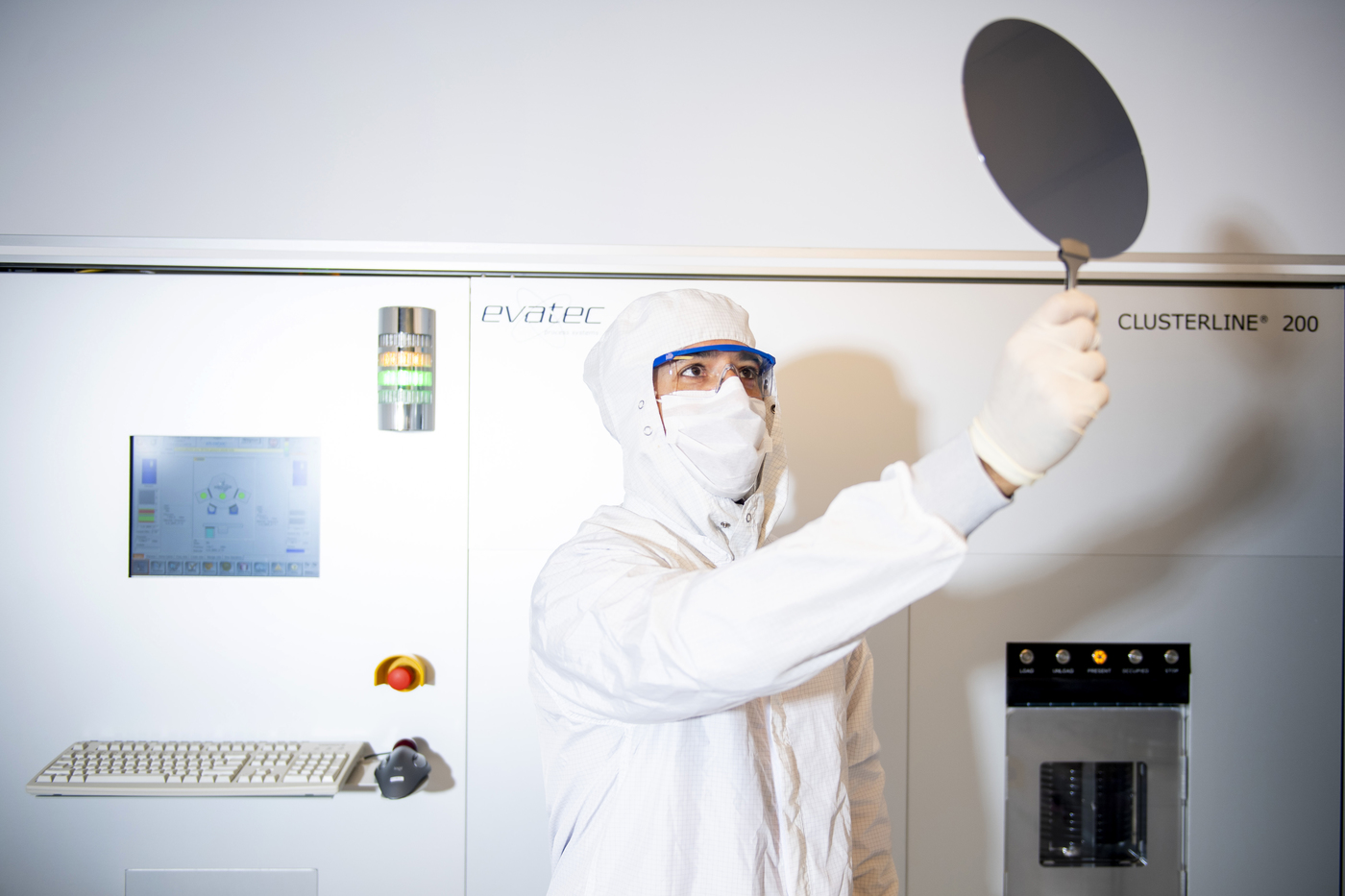 professor in clean room lab clothes standing in front of Evatec's equipment in lab