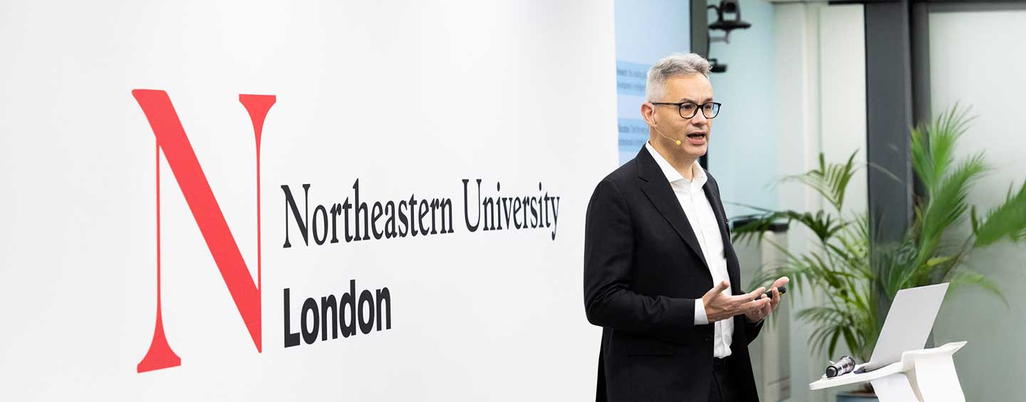 A man in a suit speaking at a podium in front of a white wall featuring the Northeastern University London logo.