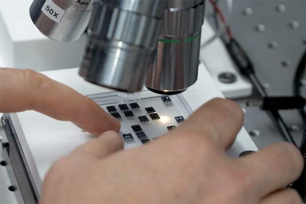 Close-up of a researcher's hands adjusting a grid of small microchips under the lens of a high-powered laboratory microscope.