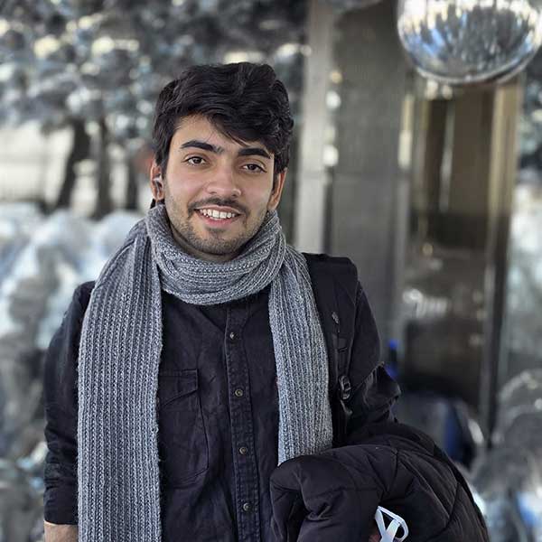 A portrait of a smiling man wearing a gray knit scarf and a dark shirt, standing outdoors with a blurred background.