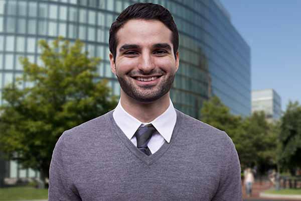 man with sweater and tie on smiling facing camera with building in background