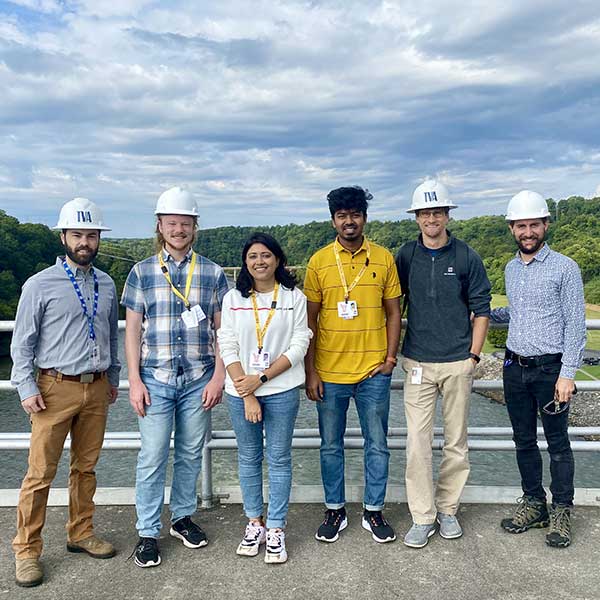 A group of six people wearing hard hats and safety lanyards standing on a bridge or dam with a river and forested hills in the background.