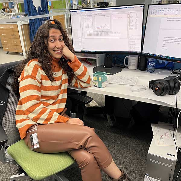 A smiling student in an orange-striped sweater sitting at a dual-monitor computer workstation in a laboratory environment.