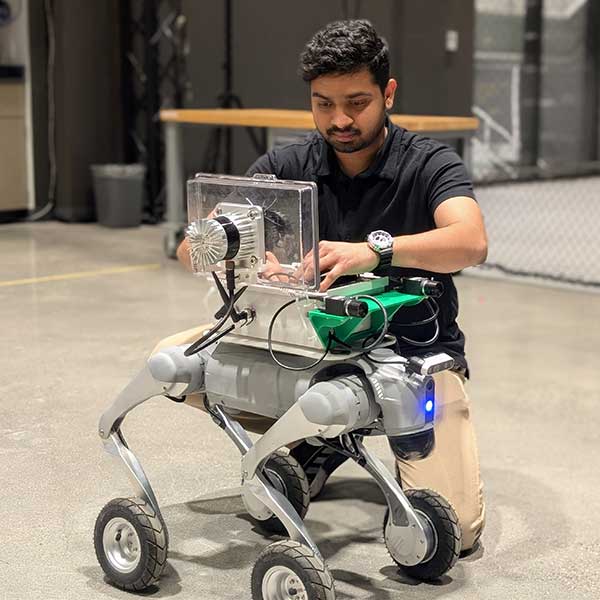 A student kneeling on a lab floor to adjust the internal components of a four-wheeled robotic platform.