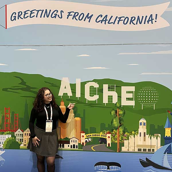A student smiles and points at a colorful "Greetings from California" mural featuring AIChE branding and California landmarks.