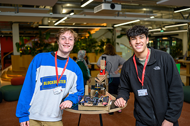 Two students smile at the camera standing by their project