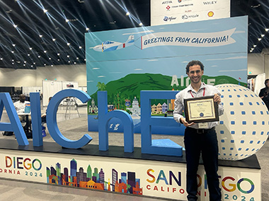 Dominic Pizzarella, chemical engineering student, standing in front of large AIChE sign holding an award.