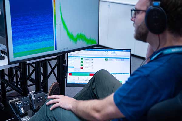 Researcher in a blue shirt wearing headphones while monitoring data on two computer screens