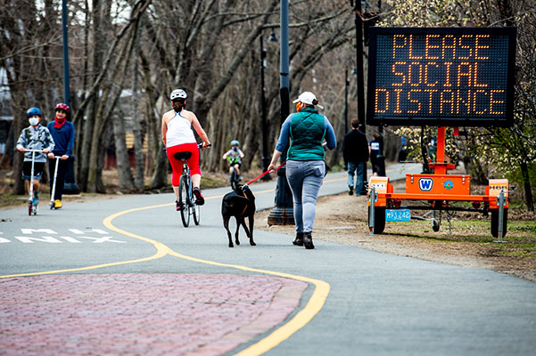 people walking and riding bike on walkway with please social distance digital sign