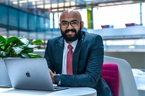 male in business suit looking at camera while sitting at table with laptop in the interdisciplinary science and engineering complex at Northeastern